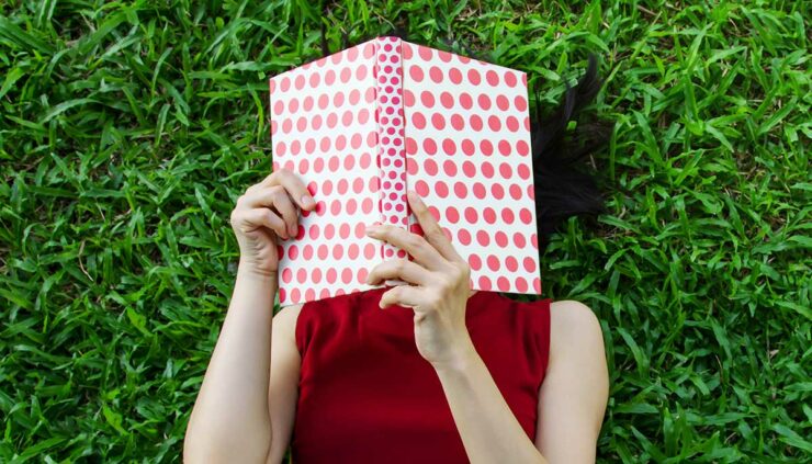 A woman in a red dress holds a book over her face as she lies back on bright green grass