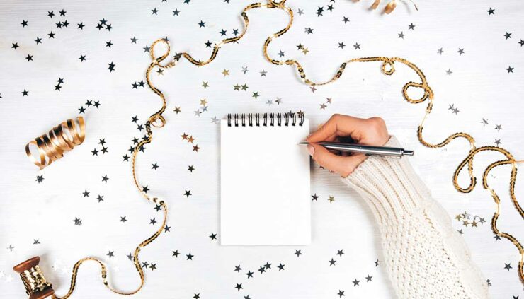 A woman's hand holds a pen, ready to write in a blank notebook surrounded by gold confetti