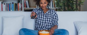 A woman holding a bowl of popcorn uses a remote control to change the channel