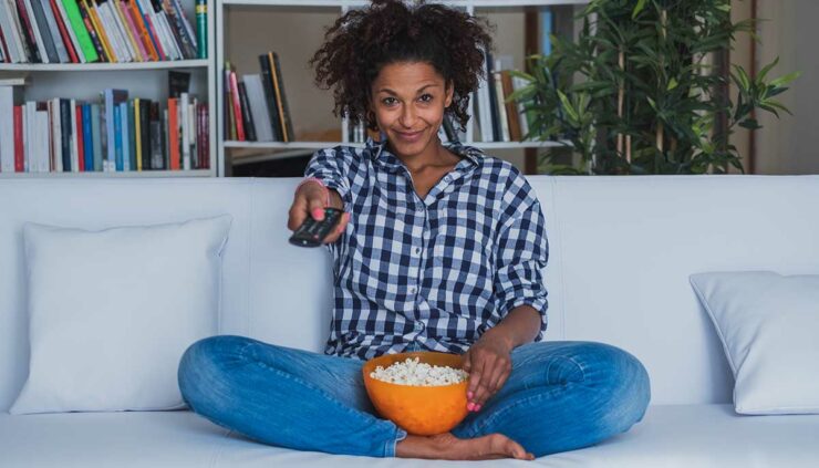 A woman holding a bowl of popcorn uses a remote control to change the channel