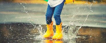 A child wearing bright yellow boots and holding a rainbow umbrella splashes in a puddle