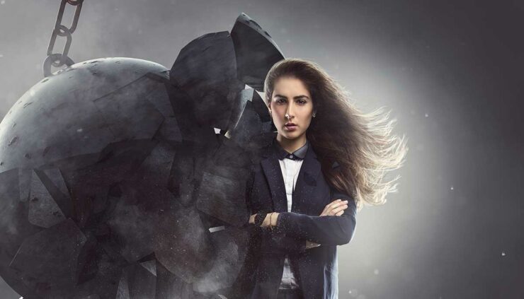 A long-haired woman in a suit stands stoically as a wrecking ball breaks against her shoulder