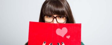 an introverted woman peeks from behind a red book with a heart on the cover