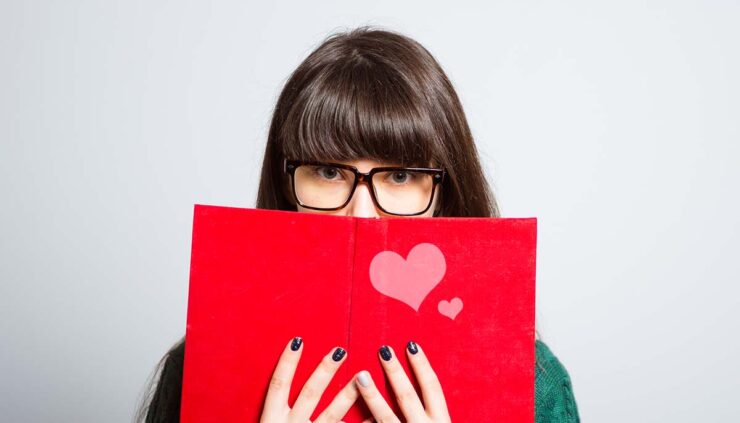 an introverted woman peeks from behind a red book with a heart on the cover
