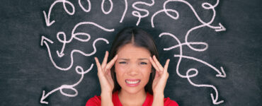 An asian woman in a red shirt stands in front of a chalkboard depicting confusing arrows sprouting out of her head.