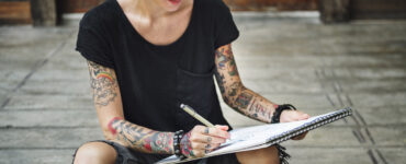 A tattoed woman in black wearing sunglasses draws on a sketch pad.