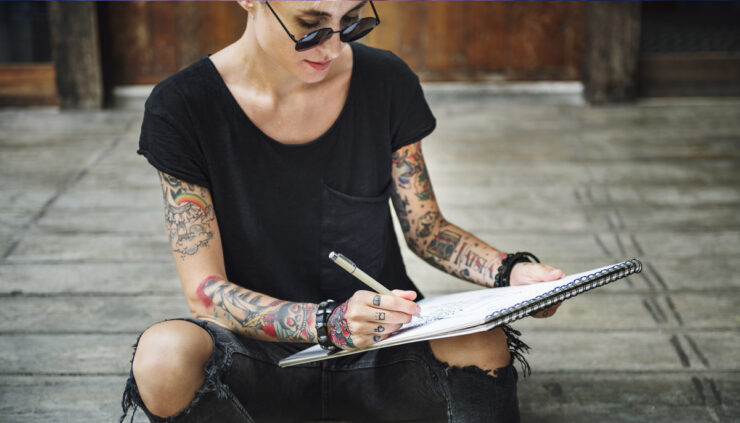A tattoed woman in black wearing sunglasses draws on a sketch pad.