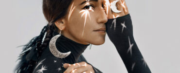 A beautiful young brunette woman in a dark turtleneck and wearing large crescent moon earrings and ring has stars reflecting off of her.