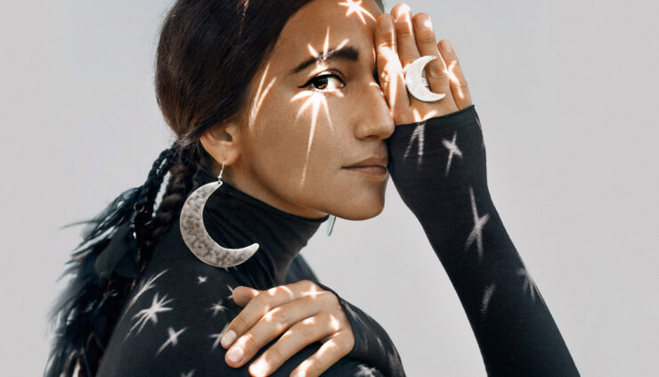 A beautiful young brunette woman in a dark turtleneck and wearing large crescent moon earrings and ring has stars reflecting off of her.