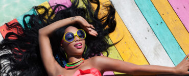 An African American woman wearing sunglasses and a brightly colored swimsuit lays in the sun on a multicolored boardwalk.