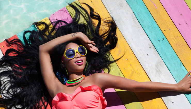 An African American woman wearing sunglasses and a brightly colored swimsuit lays in the sun on a multicolored boardwalk.