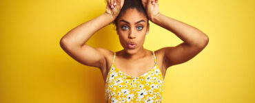 African american woman wearing casual floral dress standing over isolated yellow background doing funny gesture with finger over head as bull horns.