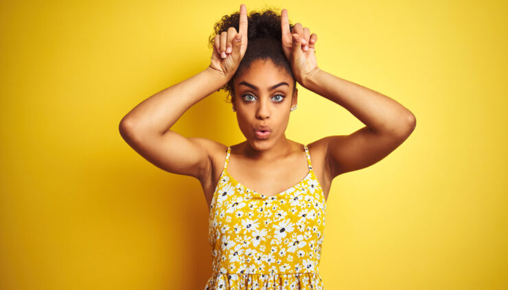 African american woman wearing casual floral dress standing over isolated yellow background doing funny gesture with finger over head as bull horns.