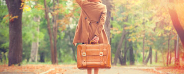 A woman in a dress, trench coat, and hat holds a suitcase behind her as she looks down a tree-lined road.