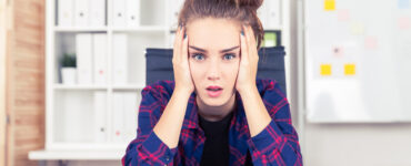 A young girl in a plaid shirt and messy bun holds her head in her hands in frustration.