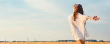 A blonde woman in a flowy white dress stands in an open field with her arms outstretched.