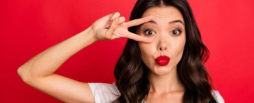 Beautiful brunette woman on a red background wears red lipstick and holds a v-sign around her eye.