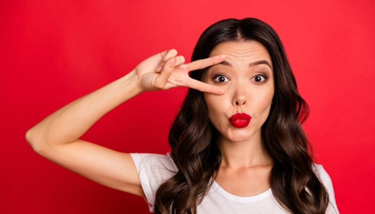 Beautiful brunette woman on a red background wears red lipstick and holds a v-sign around her eye.