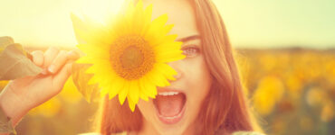 Smiling woman in a sun drenched field holds a sunflower over one eye.