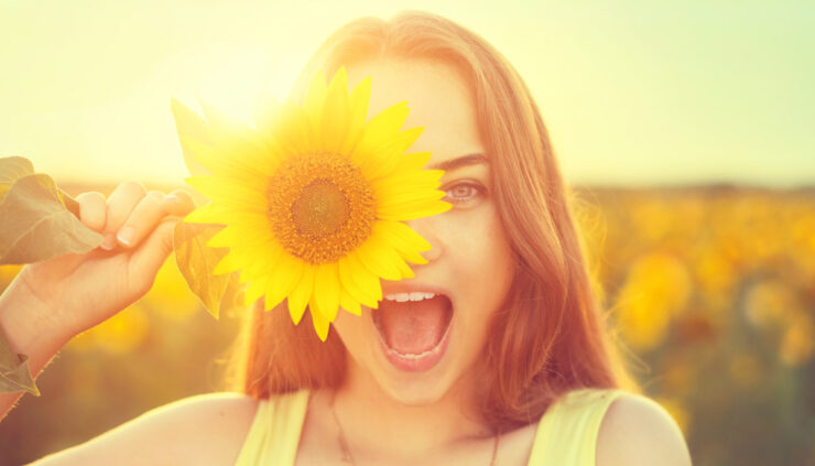 Smiling woman in a sun drenched field holds a sunflower over one eye.