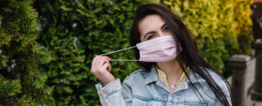 A brunette woman puts on a mask while walking in a park.