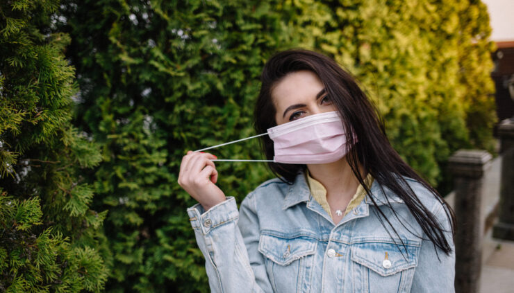 A brunette woman puts on a mask while walking in a park.