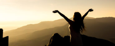 Silhouette of a woman standing on a mountain top with her arms upraised.