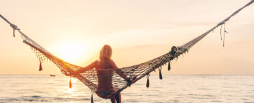 A woman in a hammock over the ocean stares off toward the sunset.