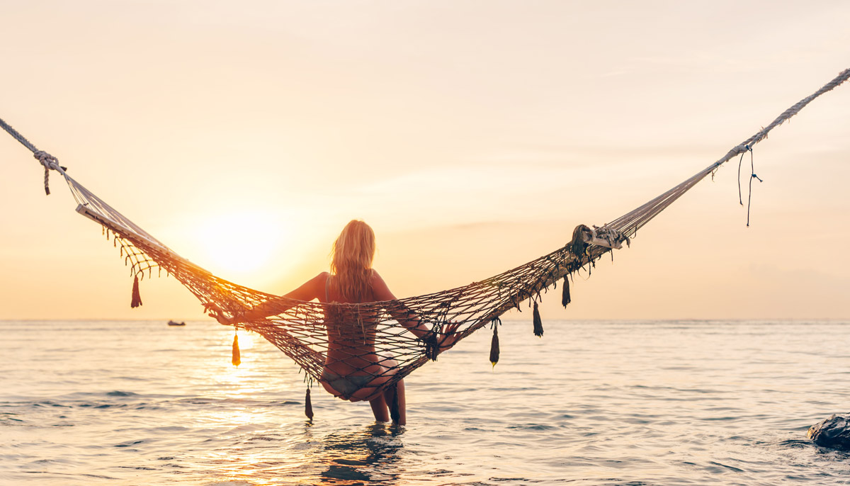 A woman in a hammock over the ocean stares off toward the sunset.