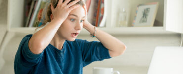 A frazzled blond woman stares at her laptop in disbelief.