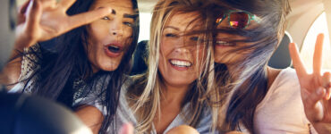 Three windswept girls riding in the backseat of a car flash peace signs at the camera.