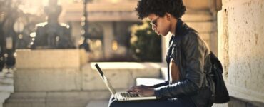 Beautiful black woman sits on the steps of a city building with a laptop.