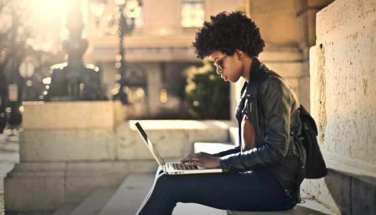 Beautiful black woman sits on the steps of a city building with a laptop.