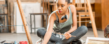 African woman surrounded by easels in art studio paints on the floor.