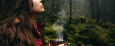 A woman in the woods holds a steaming cup of coffee.