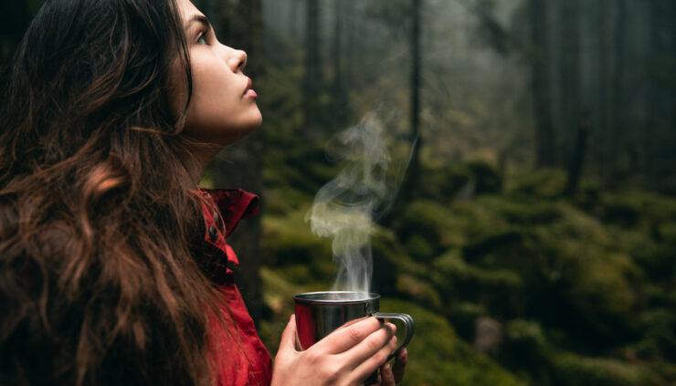 A woman in the woods holds a steaming cup of coffee.
