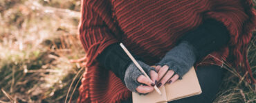 A woman in a sweater cape journals in a field.