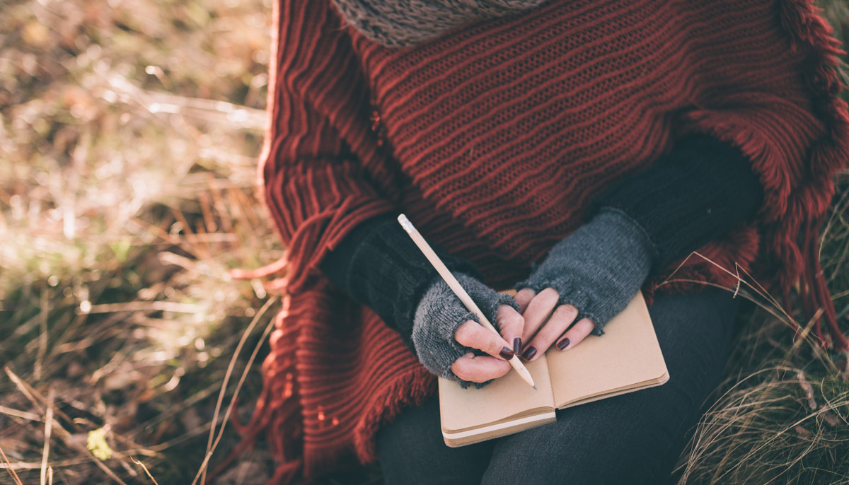 A woman in a sweater cape journals in a field.