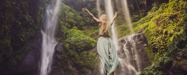 A woman in a turquoise dress standing on a cliff near a waterfall with her arms raised.